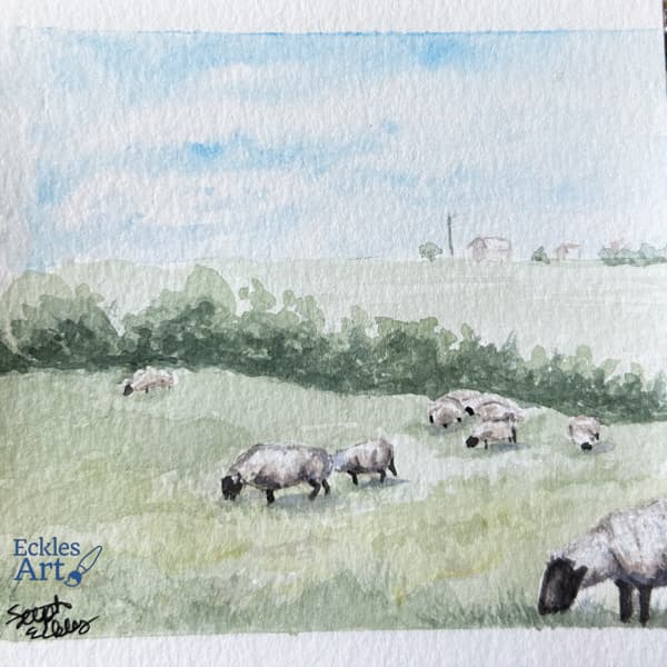 A herd of sheep scattered among a soft, pale green grass field all have heads lowered grazing. A treeline separates the foreground and background, and there is the hint of a couple of buildings beyond the field. Whispy clouds are just visible in a light blue sky.