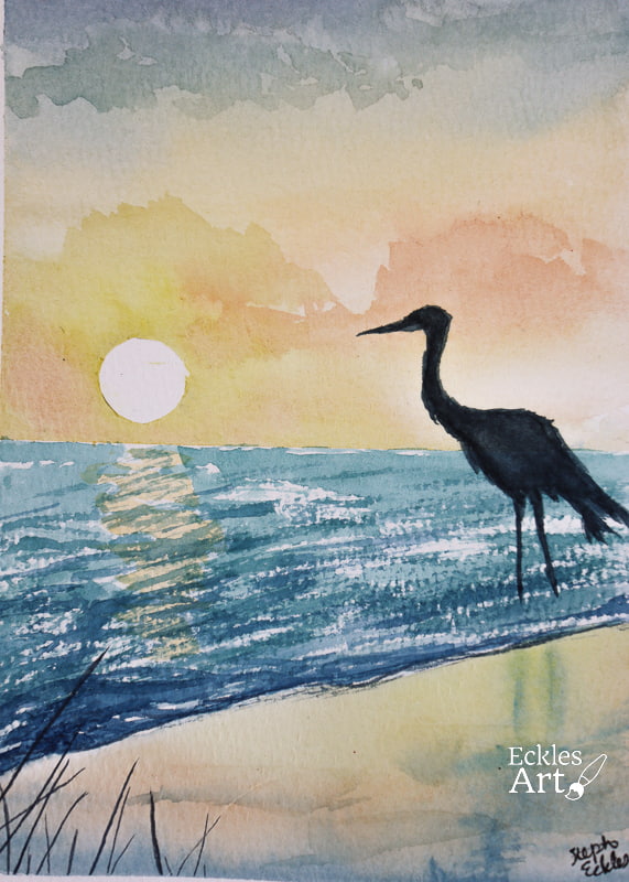 A graceful heron is silhouetted against a stunning sunset. The sky is filled with a rich gradient of warm colors, transitioning from deep oranges and pinks to yellows and blues, creating a tranquil and atmospheric backdrop. The heron, standing in shallow water just off the beach, has an elegant posture with its long legs and curved neck, emphasizing its regal presence. Reflections of the sunset hues on the water add depth and serenity to the scene.